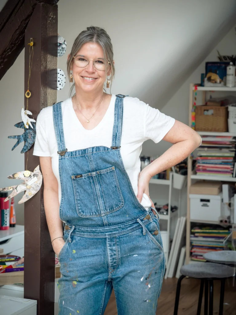 Künstlerin Stephanie Mund, mit Latzhose und weißem T-Shirt bekleidet, steht in ihrem Kunstatelier